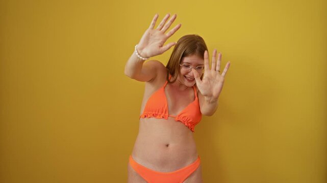 Young woman, blonde and afraid in bikini, hand raised in fear, stopping in terror, shocked expression, panic shouting over isolated yellow background