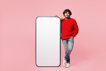 Full body young fun Indian man he wear red sweater casual clothes big huge blank screen mobile cell phone smartphone with area look camera isolated on plain pink background studio. Lifestyle concept.