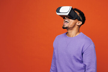 Young amazed happy man of African American ethnicity wear purple sweatshirt casual clothes watching in vr headset pc gadget isolated on plain red orange background studio portrait. Lifestyle concept.