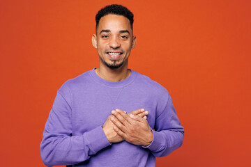 Young happy man of African American ethnicity wears purple sweatshirt casual clothes put folded hands on heart look camera isolated on plain red orange background studio portrait. Lifestyle concept.