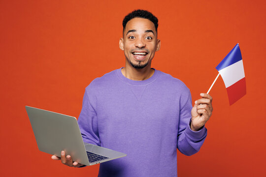 Young smart IT man of African American ethnicity wear purple sweatshirt casual clothes hold French flag use work on laptop pc computer isolated on plain red orange background studio Lifestyle concept