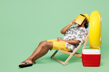 Full body side view young Indian man wear beach shirt hat rest near hotel pool sit in deckchair read book cover face sleep isolated on plain green color background Summer vacation sea sun tan concept