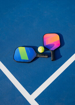 Two pickleball paddles and ball on blue outdoor court on sunny day.