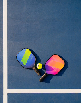 Two pickleball paddles and ball on blue outdoor court on sunny day.