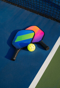 Close up of pickleball paddles and ball beside net on blue court.