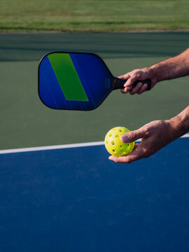 Close up of hand holding pickleball paddle and ball on outdoor court.