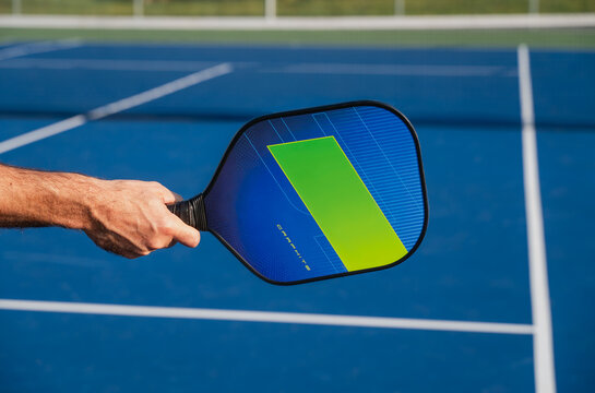 Close up of hand holding pickleball paddle on blue outdoor court.