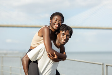 A happy African American couple shares a playful moment by the water, with the sun shining brightly. The woman smiles broadly while being playfully carried on the man back, surrounded by scenic view.
