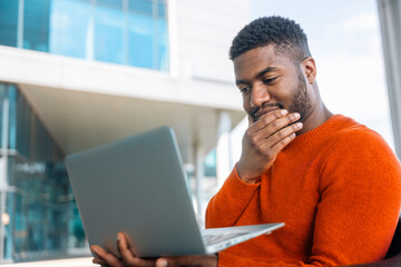 Focused man in an orange sweater engages with laptop outdoors in a modern urban setting on a sunny day