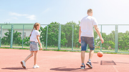 Obraz premium Girl bouncing basketball ball at outdoor court in park. Little kid practicing playing basketball with caring father. Sports leisure and weekend. Family time. Basketball game