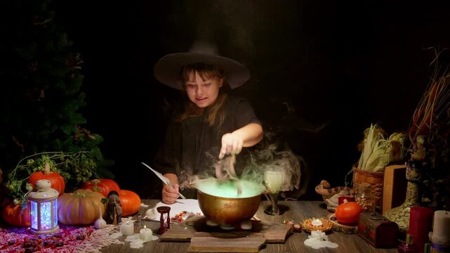 Halloween, smiling little cute girl dressed as witch in witch's hat brews a glowing magic potion in cauldron on coals. Magic wand, сandles, pumpkins, ingridients, fog smoke, magic, mystical atmosphere