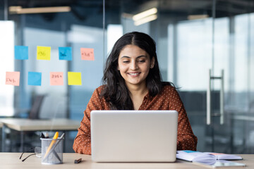 Fototapeta premium Smiling woman focused on laptop in bright office with glass wall. Workspace decorated with sticky notes, conveying creativity and organization. Atmosphere of productivity and professionalism.