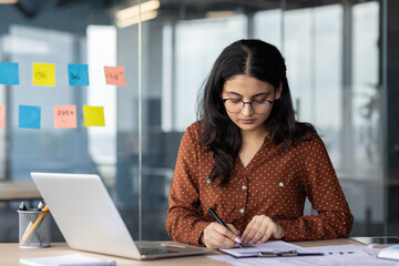 Young professional woman in office concentrating on paperwork, surrounded by laptop, and modern...