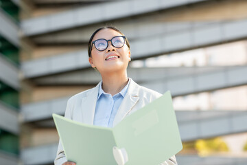 Asian woman dressed in business attire looks up with a smile while holding a folder outside a contemporary building. She appears confident and engaged, enjoying her surroundings.