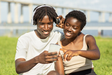 A young black couple sitting on the grass shares a smartphone while listening to music. They are smiling and enjoying each other's company on a bright, sunny day by the water.