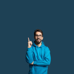 Fototapeta premium A cheerful man wearing a blue hoodie stands in a studio, smiling and pointing upward at copy space with one finger. His glasses add a playful touch while he exudes positive energy.