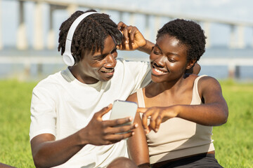 A young African American couple sits on the grass near a bridge, sharing a moment as they listen to music. They smile and laugh while looking at a smartphone, enjoying their time together.