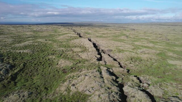 Aerial view of a large crack in the ground between two tectonic plates in a volcanic rift zone in Iceland. The landscape shows the distinct separation of the plates and the rugged terrain.