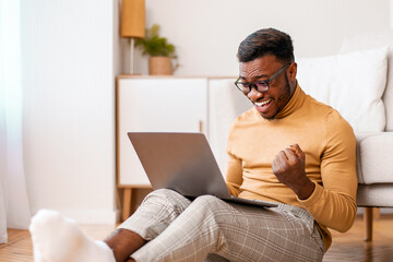 Big Luck. Excited Black Guy Using Laptop Shaking Fists Celebrating Success Sitting On Floor At Home. Selective Focus