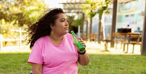 A woman sits on the grass in a park, basking in the sunlight while sipping from a green beverage. Surrounding her are trees and benches, creating a relaxing environment.