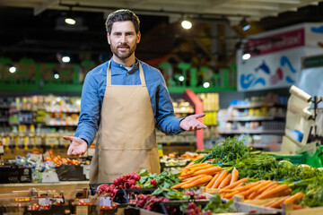 Grocery store employee in casual shirt and apron stands behind fresh produce display. Carrots and radishes neatly arranged in supermarket. Inviting atmosphere for customers