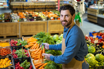 Grocery worker arranging fresh vegetables at supermarket, smiling while handling carrots. Surrounded by vibrant produce like tomatoes, radishes, and lettuce. Represents healthy eating