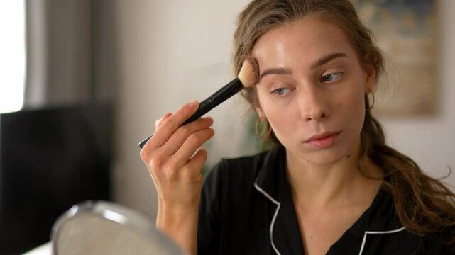 Young woman doing makeup, looking at mirror sitting in cozy room. Beautiful woman making herself beautiful indoors. Beauty, makeup concept.