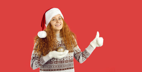 Cheerful optimistic ginger hair woman in Santa hat and mittens advertises December sale holding ball, showing thumbs up, smiling and wishing Merry Christmas and Happy New Year on red studio background