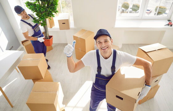 Smiling man mover captured in a portrait, confidently holding a cardboard box during an office or home relocation. With a thumbs up gesture, he signifies the success and reliability of moving service.