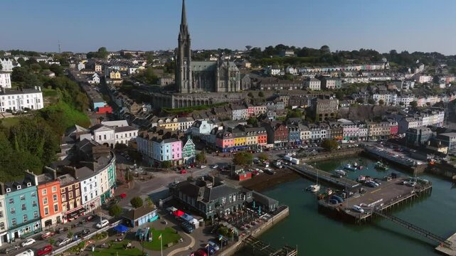 Cobh, County Cork, Ireland, September 2024. Drone circles clockwise with St. Colman's Cathedral surrounded by vibrant buildings on West Beach Promenade with a wide Seafront view in the morning light.