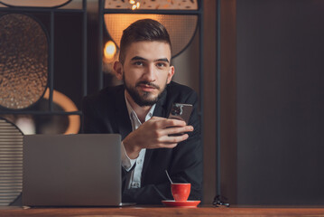 Portrait of young happy business man sitting at his desk in the modern office and using a phone.