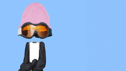 Hands Wearing Ski Gloves Are Holding Smartphone With Blank Screen Near Ski Goggles And Beanie On Blue Background With Copy Space.