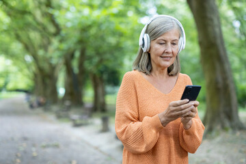 Mature woman wearing headphones using smartphone in park. She looks content, enjoying outdoor...