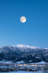  A full moon rises over a snow-covered mountain range, with a town visible in the foreground and snowy peaks in the background