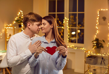 Loving couple celebrates Valentine Day at home, holding a red heart gift and embracing. The holiday captures love, affection, and togetherness, creating a joyful and romantic atmosphere.