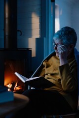 Senior woman, wrapped in warm knitted plaid, relaxing at home, sitting in chair by fireplace and reading a book