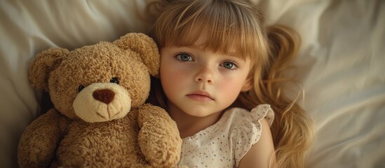 A little girl with blonde hair lies in bed with a teddy bear, looking thoughtfully at the camera.