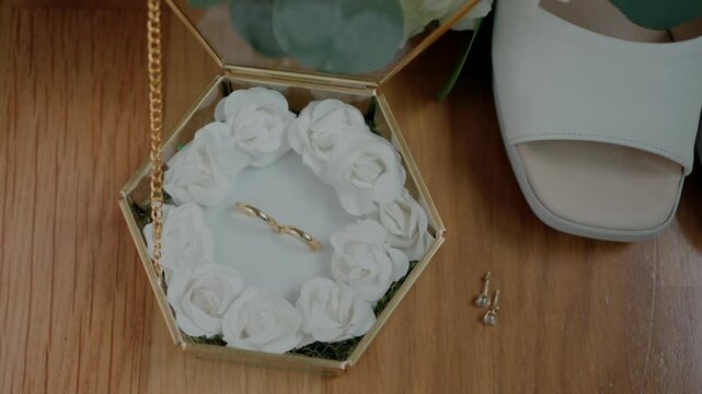 Golden wedding bands on white flowers in hexagonal box with shoe, earrings nearby.