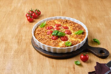 Frittata, egg casserole with cherry tomatoes, cheese and cornmeal in a light ceramic dish on a wooden background. Egg dishes.