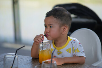 A two-year old boy is drinking a glass of orange juice using a straw; serious expression