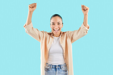 Obraz premium Portrait of a Caucasian young adult woman celebrating success and achievement, exclaiming yes and raising hands in accomplishment. She is happy and joyful, set against a blue background.