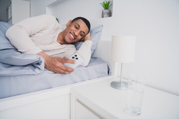 Photo of nice young man lying bed hold device wear pajama enjoy wake up modern cozy interior bedroom indoors