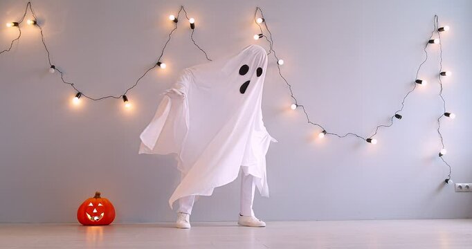 An actor in a white ghost costume dances funny in the studio. A cheerful ghost dancer with an orange pumpkin nearby. Concept idea for Halloween party, bedsheet ghost 
