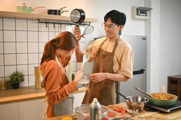 Cheerful asian young couple playfully fighting in kitchen with kitchenware