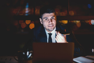 Busy Entrepreneur Using Smartphone Voice Assistant Working On Laptop In Modern Office Late At Night. Selective Focus