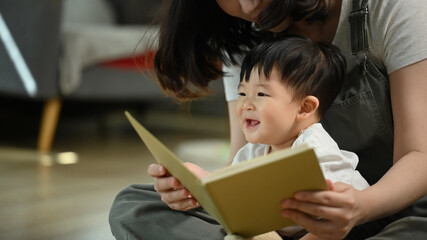Naklejka premium Mother reading book to her adorable toddler son on floor in living room. Childhood and nurturing care concept