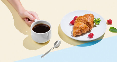 Hand holding a cup of espresso coffee on a yellow and blue background with shadow and fresh croissants with raspberry.