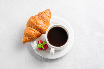 Cup of espresso coffee and croissant with fresh raspberries on a plate on a white texture  background with morning light.