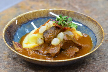 A traditional Czech dish served on a rustic plate, featuring slow-braised beef cheeks in a rich, flavorful sauce, accompanied by crispy bacon, caramelized onions, and creamy mashed potatoes.