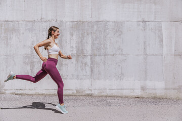 Healthy young woman in sportswear running on a sunny day. Fitness model exercising outdoors. Full length side view shot with copy space. Outdoor fitness and healthy lifestyle concept.